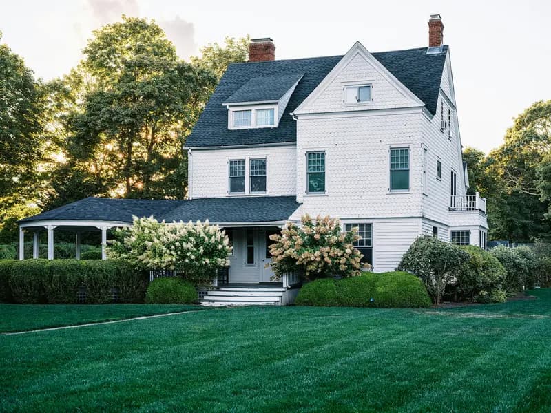 A white shingle-style house with a front porch, hydrangeas and a wide green lawn